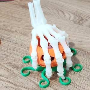 May include: A white plastic skeleton hand holding a small orange pumpkin with a green stem. The pumpkin is sitting on a green plastic base with a vine design.