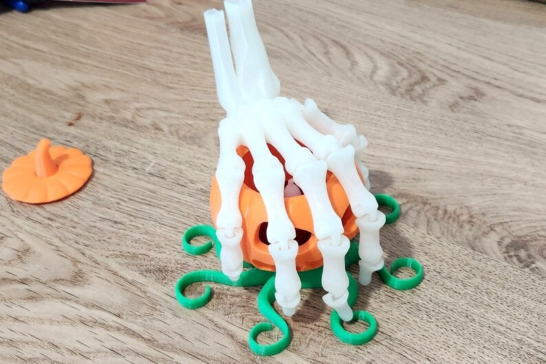 May include: A white plastic skeleton hand holding a small orange pumpkin with a green stem. The pumpkin is sitting on a green plastic base with a vine design.
