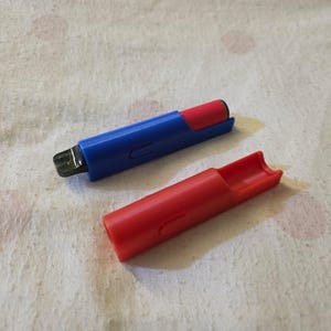 K&ouml;nnte beinhalten: Ein blau-rotes Feuerzeug mit roter Kappe. Das Feuerzeug ist blau mit rotem Oberteil und schwarzer Z&uuml;ndkomponente. Die rote Kappe ist vom Feuerzeugk&ouml;rper getrennt. Das Feuerzeug liegt auf einer wei&szlig;en Oberfl&auml;che.