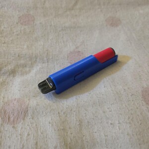 K&ouml;nnte beinhalten: Ein blau-roter Vape-Pen mit schwarzem Mundst&uuml;ck. Der Stift liegt auf einer wei&szlig;-beigen Oberfl&auml;che mit Muster. Der Vape-Pen hat eine rechteckige Form mit abgerundeten Kanten.