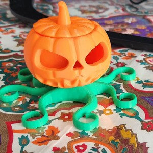 May include: A 3D printed orange pumpkin with a scary face and a green octopus base. The pumpkin has a lid and is a Halloween decoration.