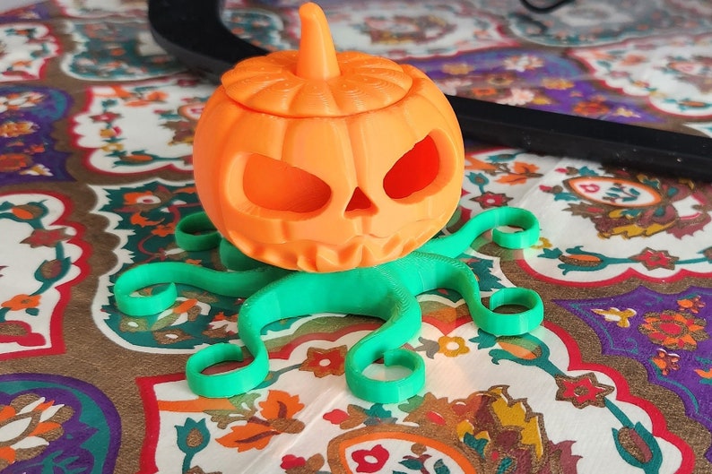 May include: A 3D printed orange pumpkin with a scary face and a green octopus base. The pumpkin has a lid and is a Halloween decoration.