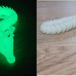 May include: A white plastic dragon figurine with a glow-in-the-dark feature. The dragon is curled up in a resting position with its head resting on its body. The dragon is shown in two images, one in a dark environment where the glow-in-the-dark feature is visible, and the other in a well-lit environment.