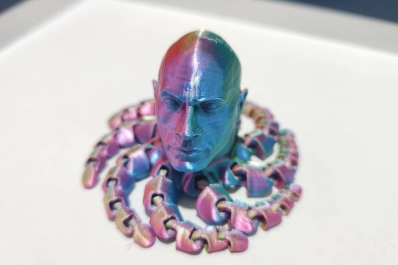 Op de afbeelding: Een 3D-geprint hoofdbeeld met een iriserende blauwe, roze en paarse afwerking. Het hoofd is omringd door een ketting van met elkaar verbonden 3D-geprinte stukken.