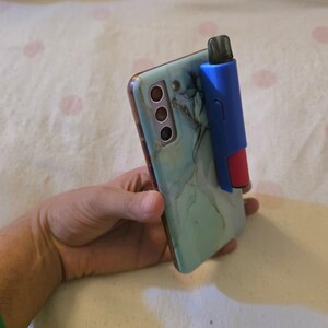 K&ouml;nnte beinhalten: Ein t&uuml;rkisfarbenes Smartphone mit einer H&uuml;lle im Marmordesign, gehalten in einer Hand. Befestigt ist ein blaues und rotes Zubeh&ouml;r, m&ouml;glicherweise ein Vape Pen-Halter. Das Telefon hat mehrere Kameralinsen.