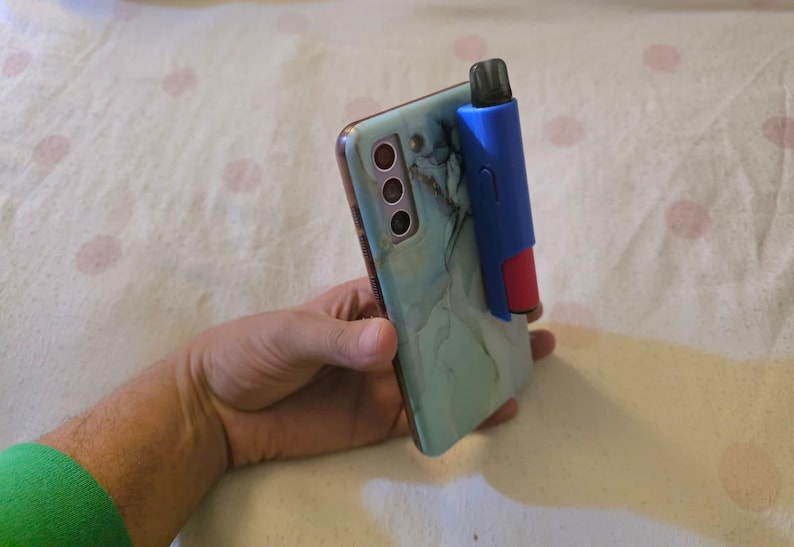 K&ouml;nnte beinhalten: Ein t&uuml;rkisfarbenes Smartphone mit einer H&uuml;lle im Marmordesign, gehalten in einer Hand. Befestigt ist ein blaues und rotes Zubeh&ouml;r, m&ouml;glicherweise ein Vape Pen-Halter. Das Telefon hat mehrere Kameralinsen.