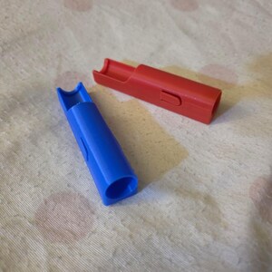 K&ouml;nnte beinhalten: Zwei Plastikpfeifen, eine blaue und eine rote, liegen auf einer strukturierten, cremefarbenen Oberfl&auml;che. Die blaue Pfeife ist zum Betrachter hin abgewinkelt, w&auml;hrend die rote Pfeife horizontal liegt. Beide haben eine zylindrische Form mit einem Schlitz.