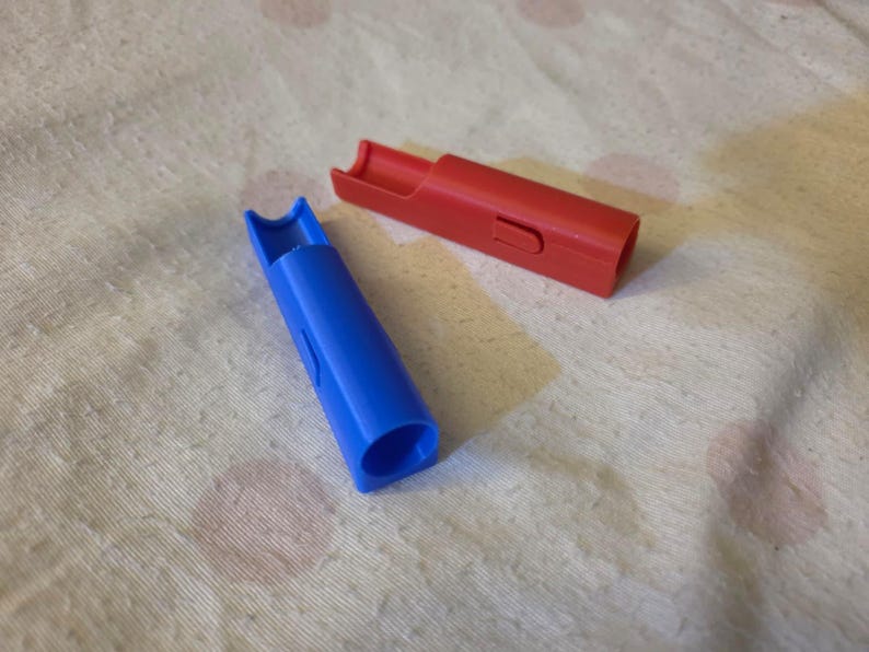 K&ouml;nnte beinhalten: Zwei Plastikpfeifen, eine blaue und eine rote, liegen auf einer strukturierten, cremefarbenen Oberfl&auml;che. Die blaue Pfeife ist zum Betrachter hin abgewinkelt, w&auml;hrend die rote Pfeife horizontal liegt. Beide haben eine zylindrische Form mit einem Schlitz.