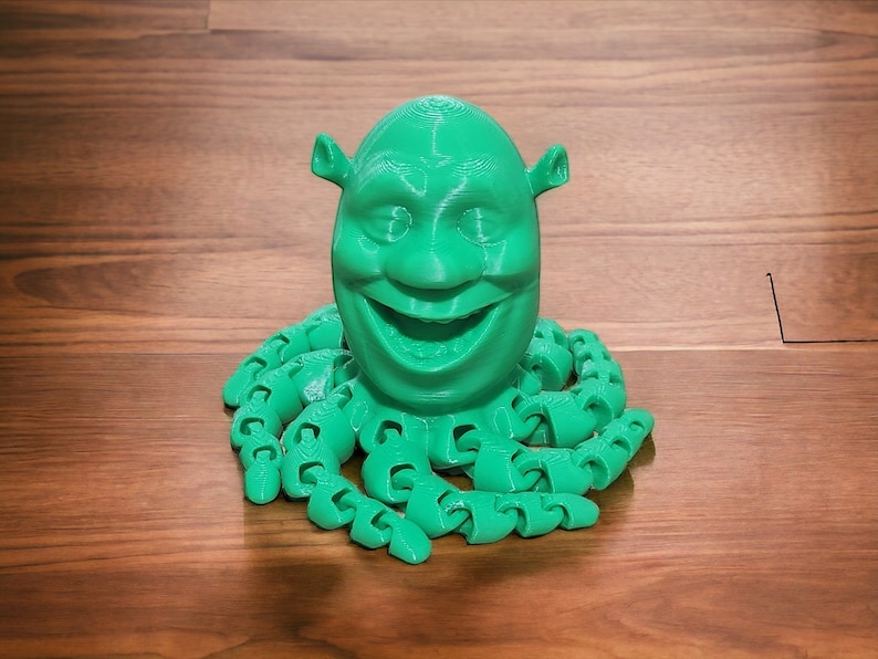 Puede incluir: Una figura impresa en 3D de color verde de la cabeza de Shrek con una expresi&oacute;n sonriente, unida a un cuerpo verde de pulpo con m&uacute;ltiples tent&aacute;culos.