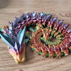 May include: A 3D printed dragon figurine with a rainbow iridescent finish. The dragon is curled up in a sleeping position with its head resting on its tail. The dragon's scales are made to look like crystals.