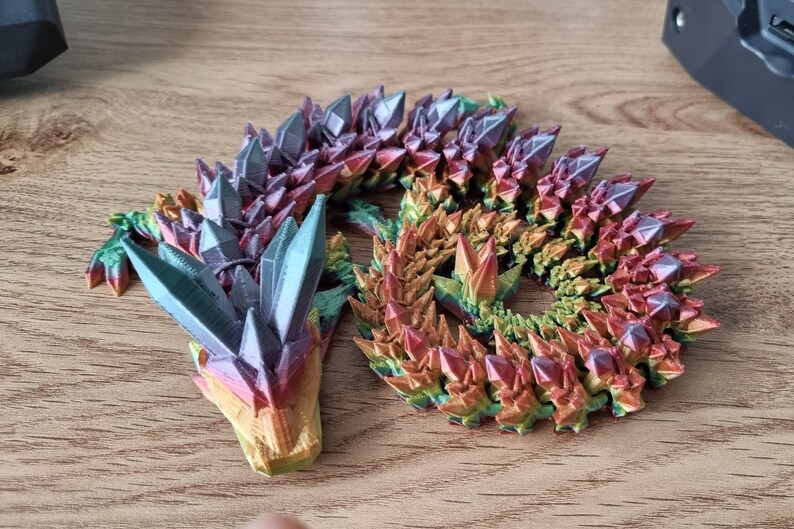 May include: A 3D printed dragon figurine with a rainbow iridescent finish. The dragon is curled up in a sleeping position with its head resting on its tail. The dragon's scales are made to look like crystals.