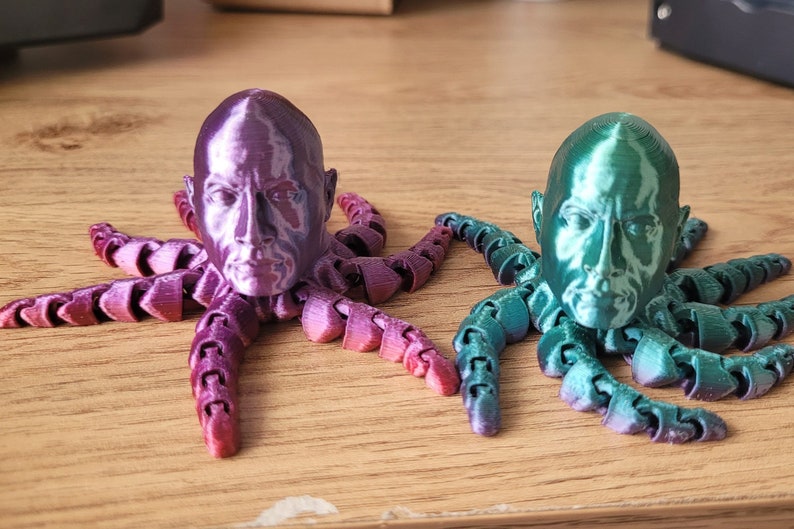 Op de afbeelding: Twee 3D-geprinte octopus-beeldjes met menselijke gezichten. De octopus links is paars en de octopus rechts is groen.