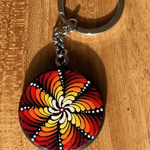 Könnte beinhalten: Ein runder Schlüsselanhänger mit einem bemalten Mandala-Design. Das Mandala zeigt ein Spiralmuster in Rot-, Orange-, Gelb- und Weißtönen auf schwarzem Hintergrund. Der Schlüsselanhänger hat einen silberfarbenen Ring und eine Kette.