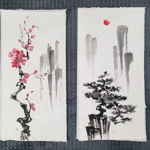 May include: Two watercolor paintings on textured paper. One features a flowering branch with pink blossoms. The other depicts a tree atop a rock formation with a red sun and gray mountains in the background. Both are in a minimalist style.