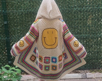 Puede incluir: Cárdigan con capucha de crochet color crema con una gran cara sonriente amarilla en la espalda y caras sonrientes más pequeñas en las mangas. El cárdigan presenta patrones cuadrados coloridos y un diseño geométrico.