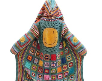 Puede incluir: Una larga chaqueta de punto de ganchillo verde con un diseño de patchwork colorido y una gran cara sonriente amarilla en la espalda. La chaqueta tiene capucha y está hecha con una variedad de hilos de colores.