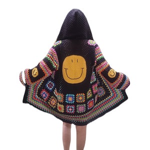 Könnte beinhalten: Ein schwarzer Häkelcardigan mit einem bunten Granny-Square-Muster und einem gelben Smiley auf der Rückseite. Der Cardigan hat eine Kapuze.