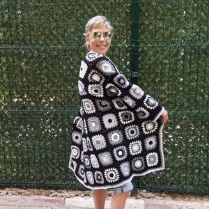 May include: A black and white crochet cardigan with a geometric pattern. The cardigan is long and has a loose fit. The person wearing the cardigan is standing in front of a green fence.