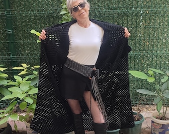 Puede incluir: Una mujer con un cárdigan de crochet negro, una camiseta blanca, una minifalda negra, medias de rejilla negras y botas negras de caña alta. Está de pie frente a una valla verde.