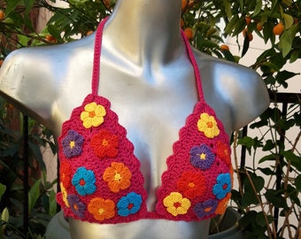 Op de afbeelding: Een handgemaakte, felroze gehaakte bikinitop met kleurrijke bloemenapplicaties. De bloemen zijn in tinten geel, oranje, blauw en paars. De top wordt getoond op een zilveren mannequin, met een achtergrond van groen gebladerte en sinaasappels.