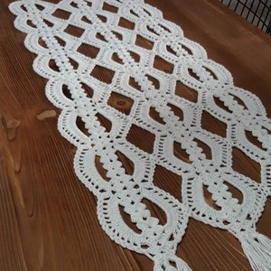 Pode incluir: Um corredor de mesa de crochê branco com um padrão de diamante e franjas. O corredor está colocado sobre uma mesa de madeira.