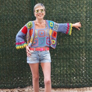 May include: A woman wearing a colorful crocheted top and matching cardigan with a bright pink belt. She is wearing denim shorts and blue shoes.