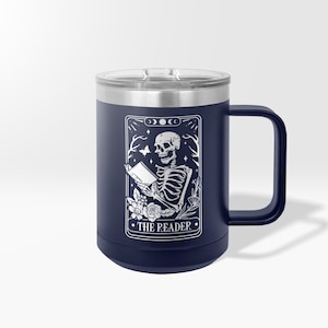 May include: A navy blue stainless steel travel mug with a white design of a skeleton reading a book. The design is surrounded by flowers and the text "THE READER".