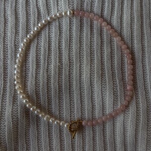 May include: A necklace featuring a combination of white pearls and pink beads. The necklace is secured with a gold-colored clasp. The jewelry is displayed on a textured, light gray surface.