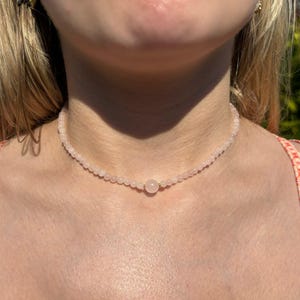 Könnte beinhalten: Eine zarte Halskette aus Rosenquarz-Perlen. Die Halskette besteht aus kleinen, runden Perlen in einem zarten Rosa, die zu einem Choker aufgereiht sind. Eine größere, einzelne Rosenquarz-Perle ist mittig angeordnet.