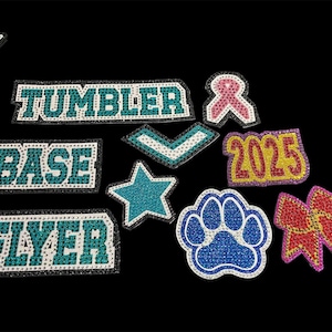 May include: A selection of rhinestone patches in various shapes and colours, including stars, a paw print, and a bow. Text patches spell out "TUMBLER", "BASE", and "FLYER". Other patches include a pink ribbon and the number "2025".