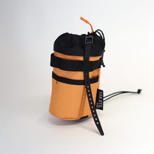 Puede incluir: Una pequeña bolsa de bicicleta naranja y negra con correas ajustables y cierre de cordón. La bolsa tiene el texto "SAH UTIL DESIGN" impreso en el lateral.