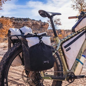 May include: A light green mountain bike with black tires and multiple attached bags for storage. The bike has a black seat and handlebars with a brown grip. The bags are black and white, and the bike is on a dirt path.