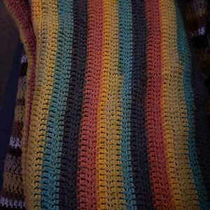 May include: A handmade crochet blanket with vertical stripes in shades of yellow, pink, blue, and gray. The blanket has a textured appearance, showcasing the individual stitches. The overall design is a blend of colors, creating a cozy and inviting aesthetic.