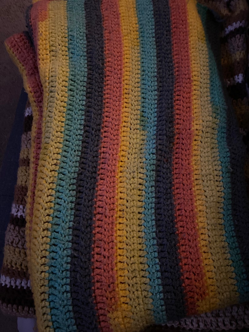 Peut inclure: Une couverture au crochet faite &agrave; la main avec des rayures verticales dans les tons de jaune, rose, bleu et gris. La couverture a un aspect textur&eacute;, mettant en valeur les points individuels. Le design g&eacute;n&eacute;ral est un m&eacute;lange de couleurs, cr&eacute;ant une esth&eacute;tique chaleureuse et accueillante.