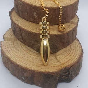 Op de afbeelding: Een gouden metalen slinger met een ketting en een decoratieve bal aan het einde. De slinger hangt aan een houten standaard.