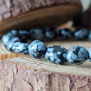 Op de afbeelding: Een close-up van een armband gemaakt van ronde, donkergrijze kralen met witte en zwarte sneeuwvlokachtige patronen. De armband ligt op een stuk getextureerd, lichtbruin hout. De kralen zijn geregen, wat een natuurlijk stenen sieradenontwerp laat zien.