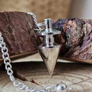 Könnte beinhalten: Ein silberfarbenes Pendel mit konischer Spitze und Kette. Das Pendel hat eine kleine, runde Kugel am Ende der Kette. Der Hintergrund ist ein Stück Holz mit natürlicher, strukturierter Oberfläche. Das Pendel ist für die Wahrsagung bestimmt.