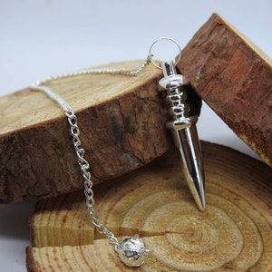 May include: A silver pendulum with a pointed tip and a chain. The pendulum is hanging from a wooden surface.