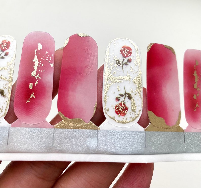 Peut inclure: Bandes de nail art avec un effet aquarelle rose et blanc, orn&eacute;es de feuilles d'or et de motifs de roses. Les d&eacute;calcomanies &agrave; ongles sont pr&eacute;sent&eacute;es sur un support argent&eacute;.