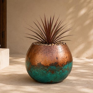 May include: A spherical metal planter featuring a hammered copper finish on the upper portion and a vibrant turquoise patina on the lower section. The planter is filled with small light-colored stones and a spiky, dark-leafed succulent plant.