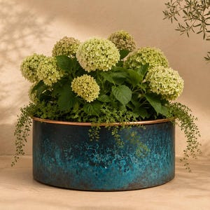 May include: A cylindrical metal planter with a dark blue and teal patina finish and a copper-colored rim. The planter is filled with a lush arrangement of pale green hydrangea blooms and trailing green foliage, creating a vibrant contrast.