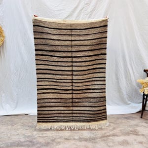May include: A rectangular rug with horizontal stripes in shades of brown and black. The rug has a cream-coloured fringe at the bottom. A straw hat and a wooden ladder are on the left. A wooden chair with pillows is on the right.