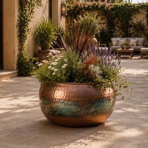 Puede incluir: Una maceta grande y redonda de cobre martillado con un acabado de pátina turquesa en la base, llena de una variedad de follaje verde, lavanda morada y pequeñas flores blancas, colocada sobre un patio de piedra en un patio exterior.