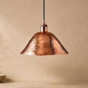 May include: A pendant light fixture featuring a wide, bell-shaped shade made of hammered copper with a metallic, reflective finish. The shade is suspended from a black cord and includes a matching copper-toned metal cap at the top connection point.