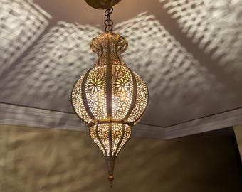 May include: An ornate, gold-coloured pendant light fixture with intricate cut-out patterns. The light is suspended from the ceiling by a chain and casts decorative shadows. The fixture has a bulb inside, illuminating the detailed design.