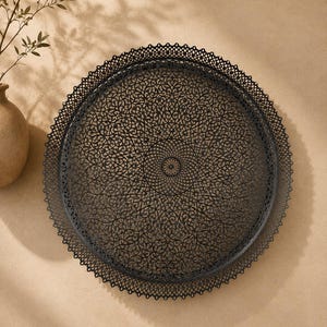 May include: A round black metal tray featuring an intricate, laser-cut filigree pattern across its surface. The tray has a delicate, scalloped lace-like border around the entire circumference, creating a detailed and openwork design throughout.