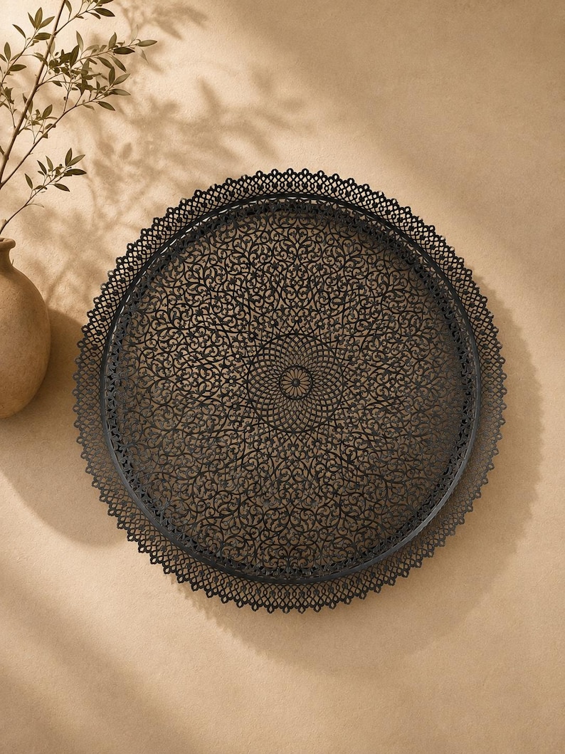May include: A round black metal tray featuring an intricate, laser-cut filigree pattern across its surface. The tray has a delicate, scalloped lace-like border around the entire circumference, creating a detailed and openwork design throughout.