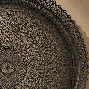 May include: A large, circular, black metal tray featuring an intricate, openwork filigree pattern with a central medallion and a scalloped, lace-like border. The dark finish has subtle bronze-toned highlights throughout the detailed metalwork.