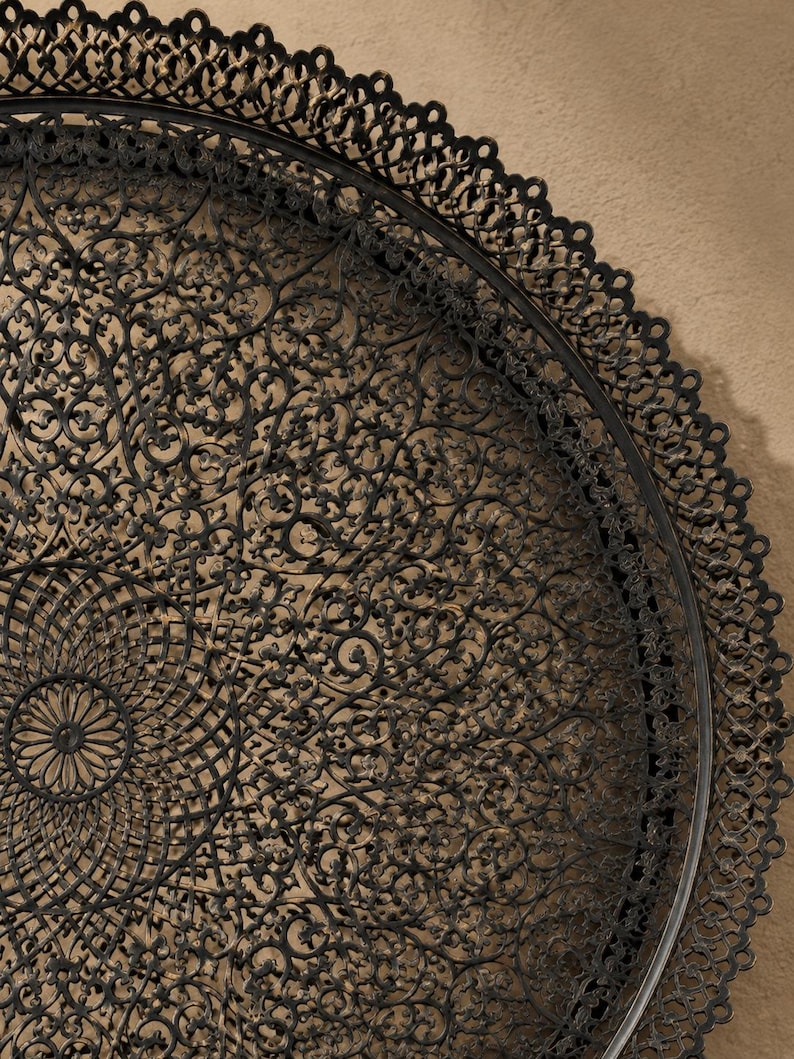 May include: A large, circular, black metal tray featuring an intricate, openwork filigree pattern with a central medallion and a scalloped, lace-like border. The dark finish has subtle bronze-toned highlights throughout the detailed metalwork.