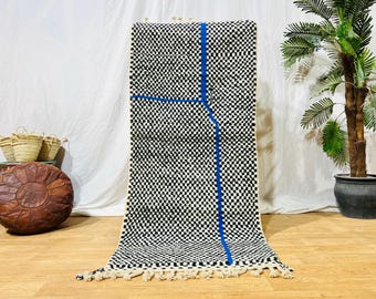 May include: A rectangular black and white checkered rug with a blue geometric design. The rug has a fringe at the bottom and is displayed on a wooden floor. A brown leather ottoman and baskets are in the background.